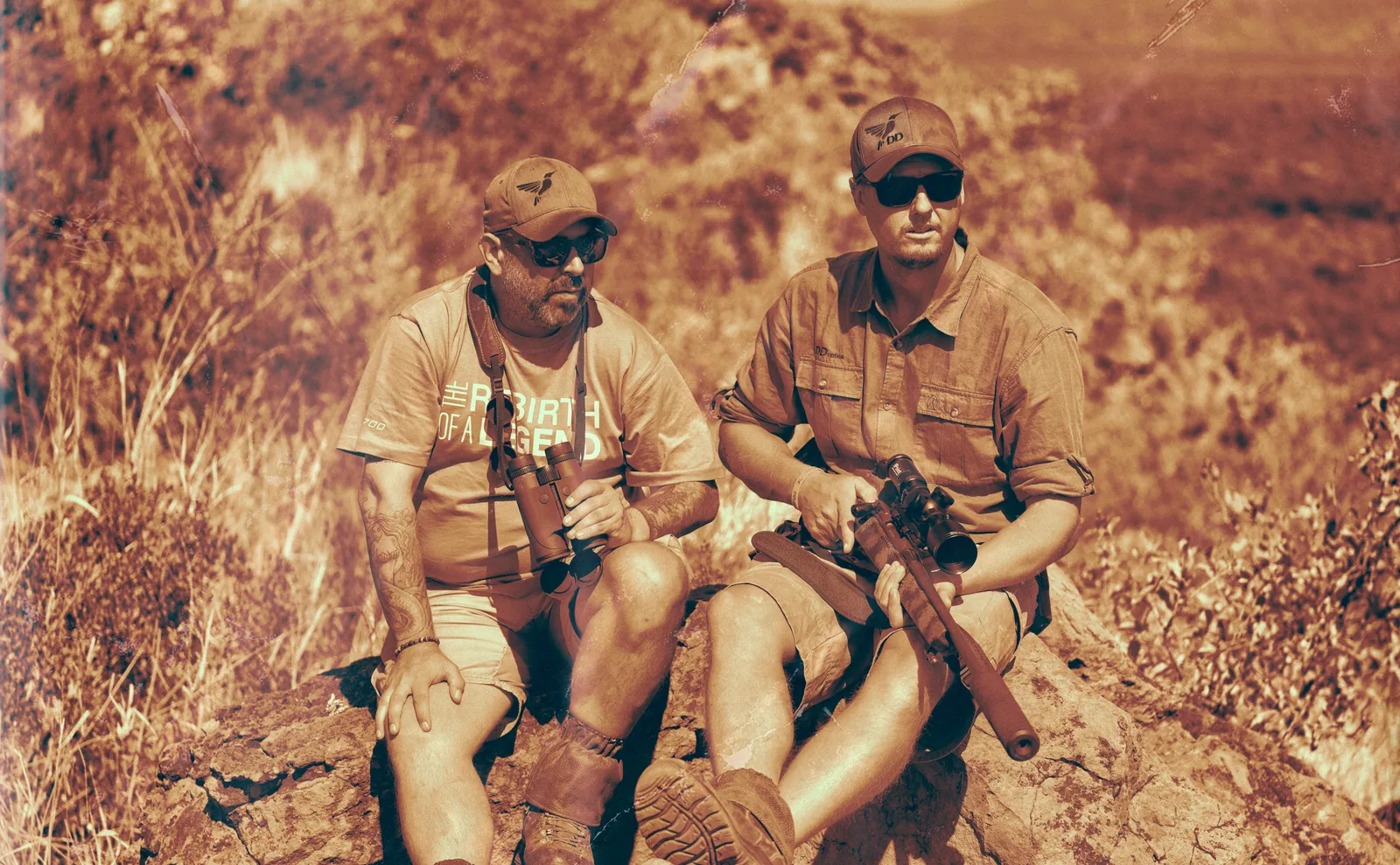 Two hunters in a rocky landscape; the hunter on the left is holding binoculars.